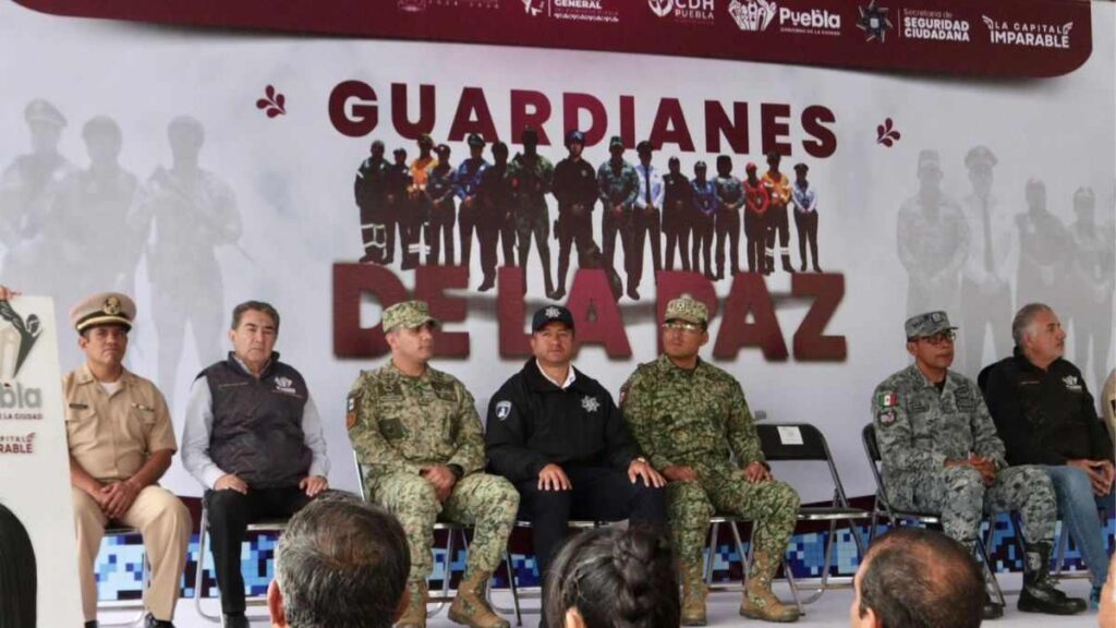 En el escenario, se encuentran sentados varios representantes de distintas instituciones de seguridad y fuerzas armadas, incluyendo elementos del Ejército, la Marina, la Policía Municipal y la Guardia Nacional.